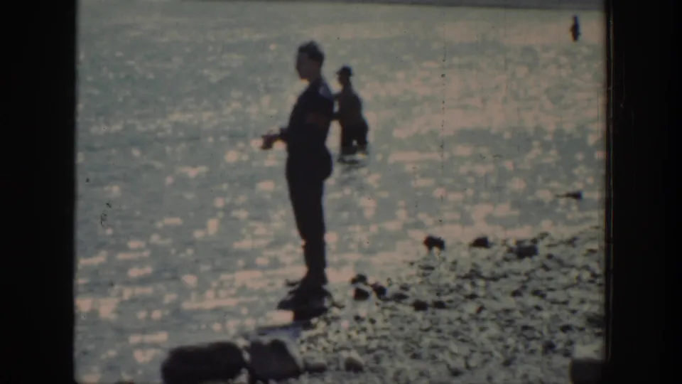 Two figures appear in a lakeside fishing scene, one near the rocky shore and one farther out in shallow water, in 1939 Wyoming home-movie footage.