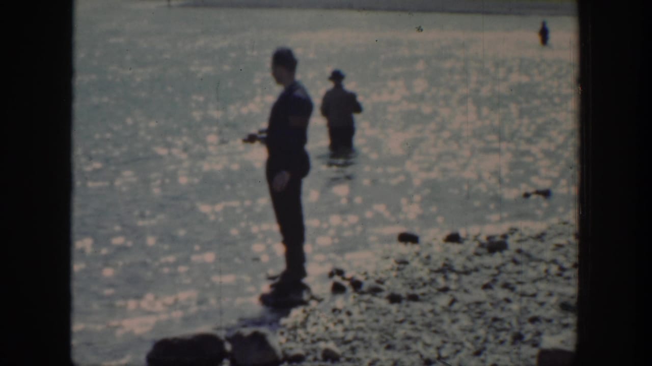 Man with fishing rod stands by the water while another figure remains farther out.