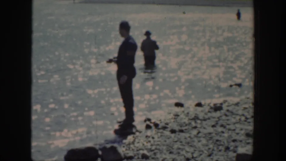 A 1939 Wyoming lakeside frame shows a man holding fishing gear near shore with a second figure standing in the water.
