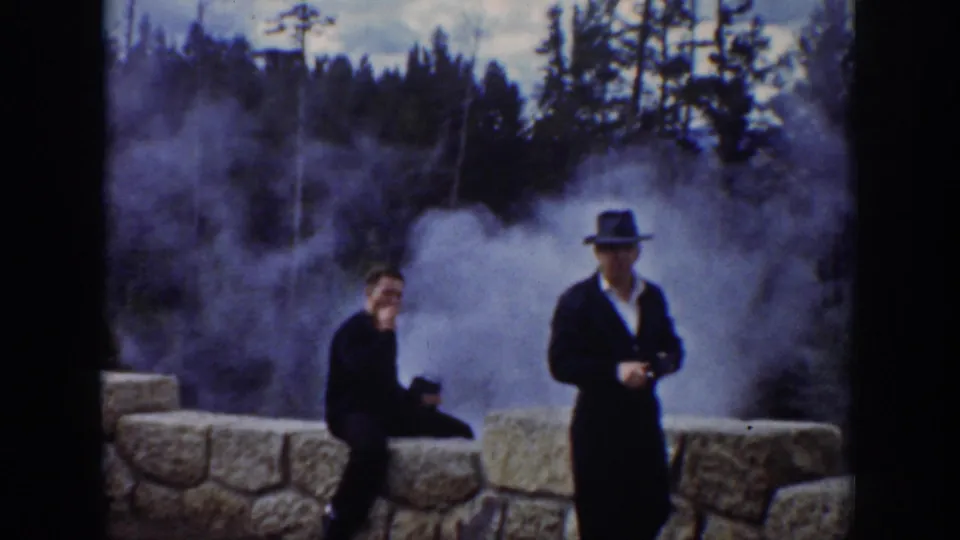 1939 Wyoming home-movie view of two adult men by a stone wall with geyser steam rising in front of pine trees.