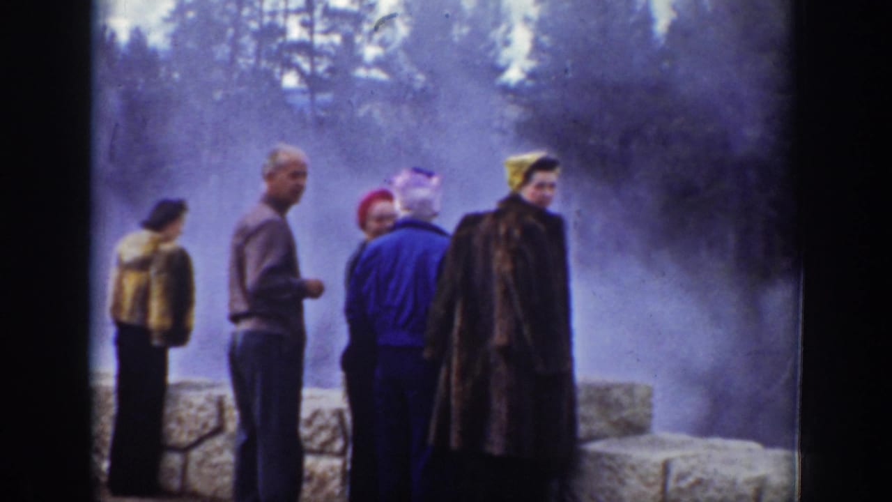 Group of adult tourists in coats and hats stands near a stone wall at a steamy Wyoming geothermal site.