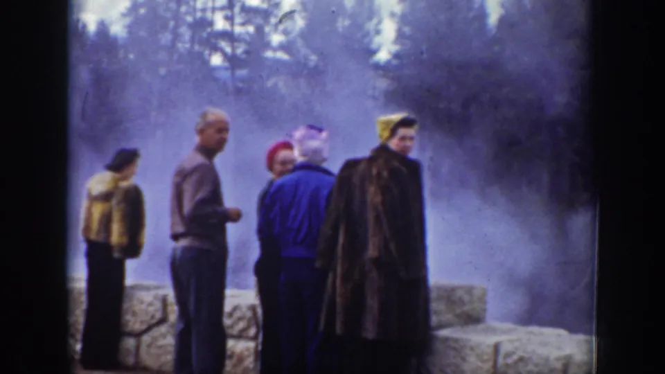 Group of adult tourists in coats and hats stands near a stone wall at a steamy Wyoming geothermal site.