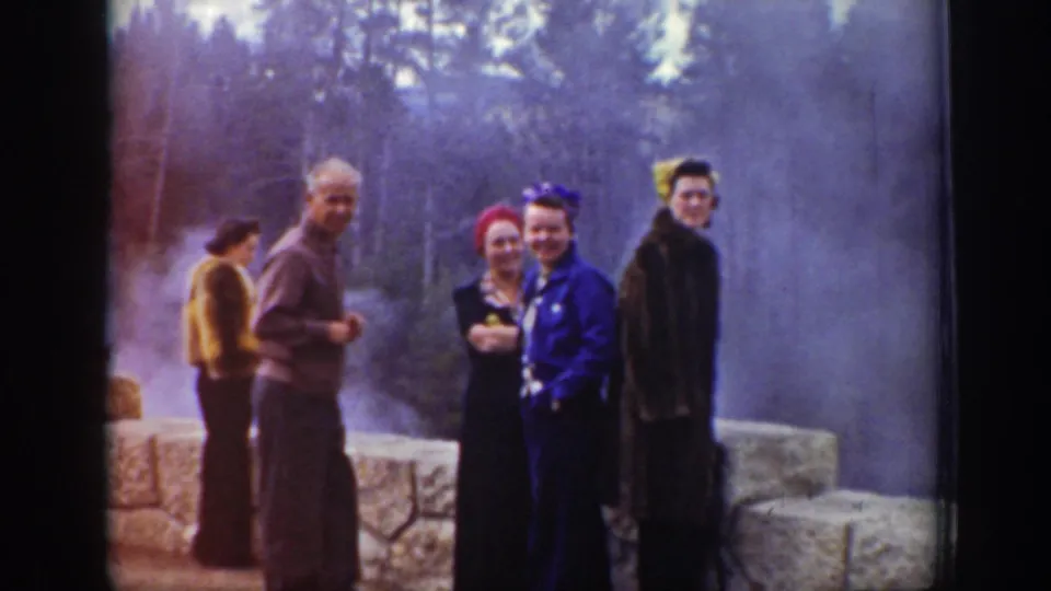 Tourists in period coats and bright hats face the camera near a steaming Wyoming geyser overlook.