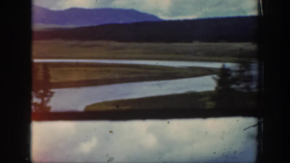 1939 Wyoming road-trip frame showing a curving river or watercourse, open valley, distant mountains, cloudy sky, and a roadside tree.