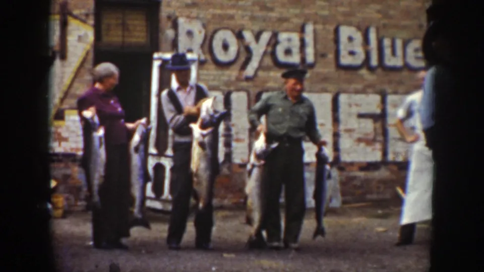 In Wyoming footage dated 1939 by metadata, three adults stand outdoors holding large fish before a brick wall with a faded Royal Blue sign.