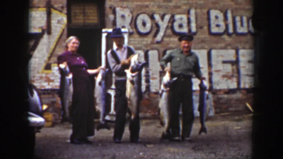 Three adults appear to smile and pose with oversized fish in the 1939 Wyoming home-movie scene, with a brick wall and faded Royal Blue sign behind them.
