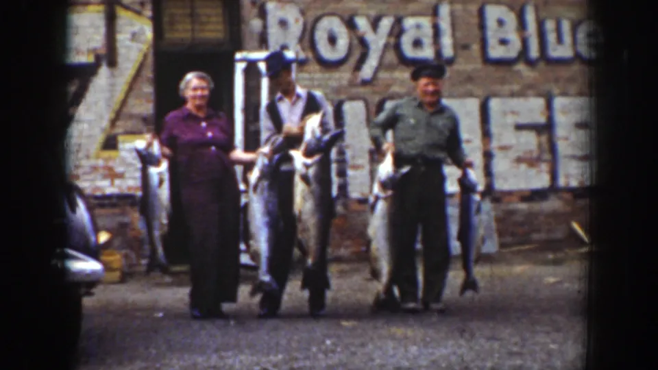 Home-movie frame from Wyoming in 1939 showing three adults smiling and holding fish before a Royal Blue wall sign.