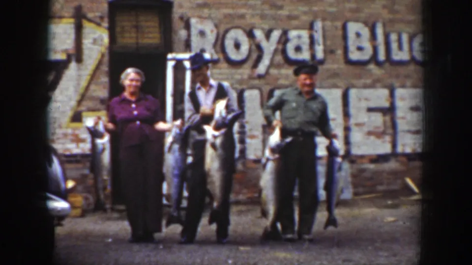 Final 1939 Wyoming frame of the group holding fish, with a brick building, faded Royal Blue sign, and partial car in view.
