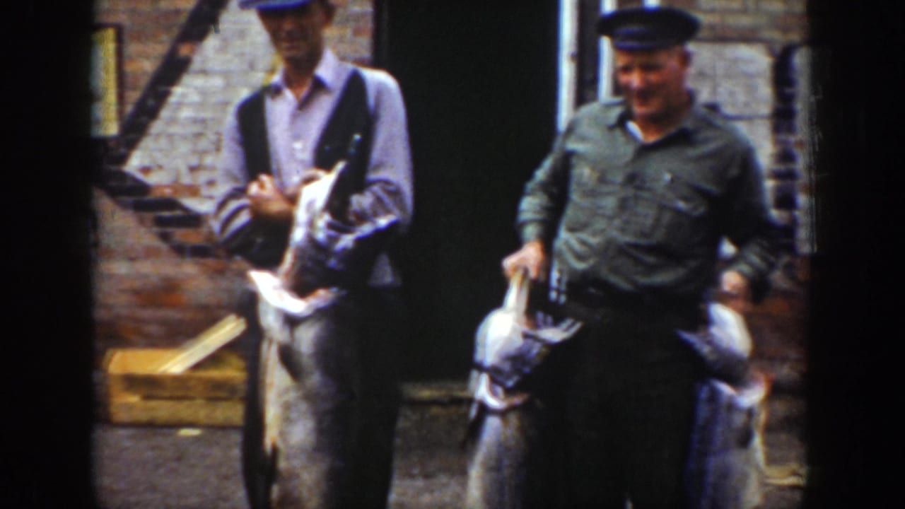 Two men in hats hold large fish in front of a brick wall and dark doorway.