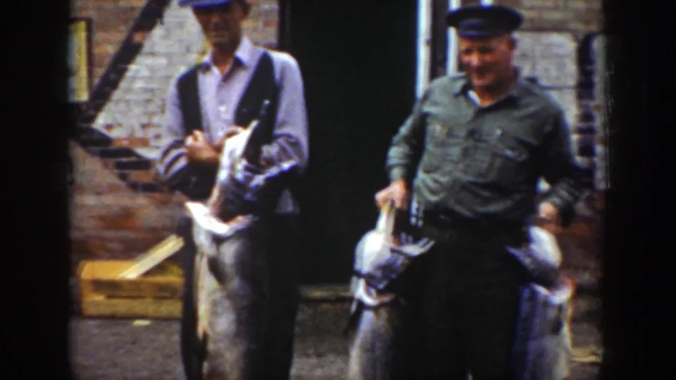 1939 Wyoming frame of two men posing with large fish outside a brick building with a faded Royal Blue wall sign nearby.