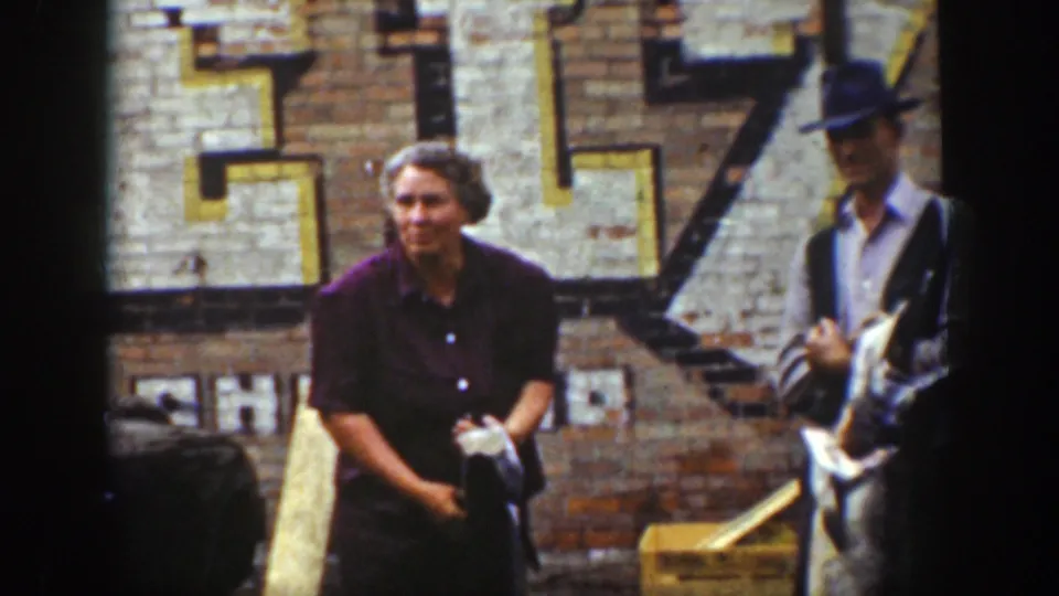 1939 Wyoming home-movie frame showing a woman in a dark dress near a man in a hat holding large fish by a brick storefront wall.