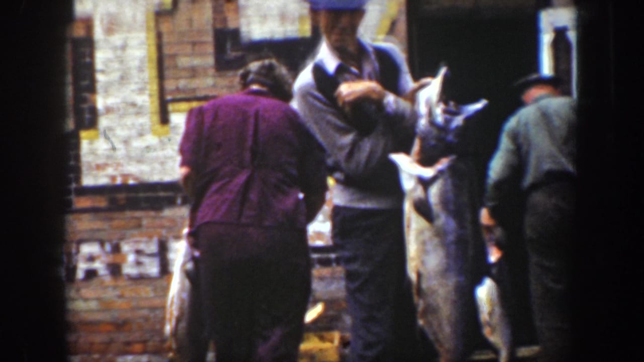 1939 Wyoming home-movie frame showing men in period clothing handling large fish near a brick wall and wooden crates.