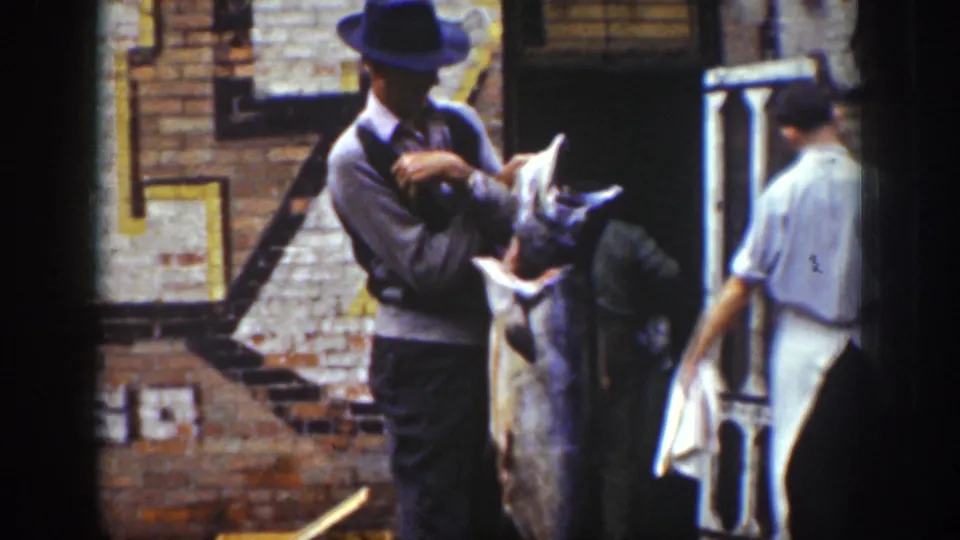 1939 Wyoming footage frame with a man in a dark hat and sweater vest holding a large fish near brick buildings and other workers.