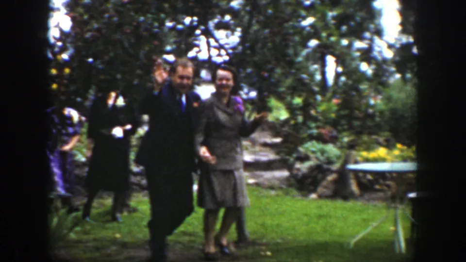 The suited man and woman smile and wave toward the camera while crossing a garden lawn in the 1939 Wyoming clip.