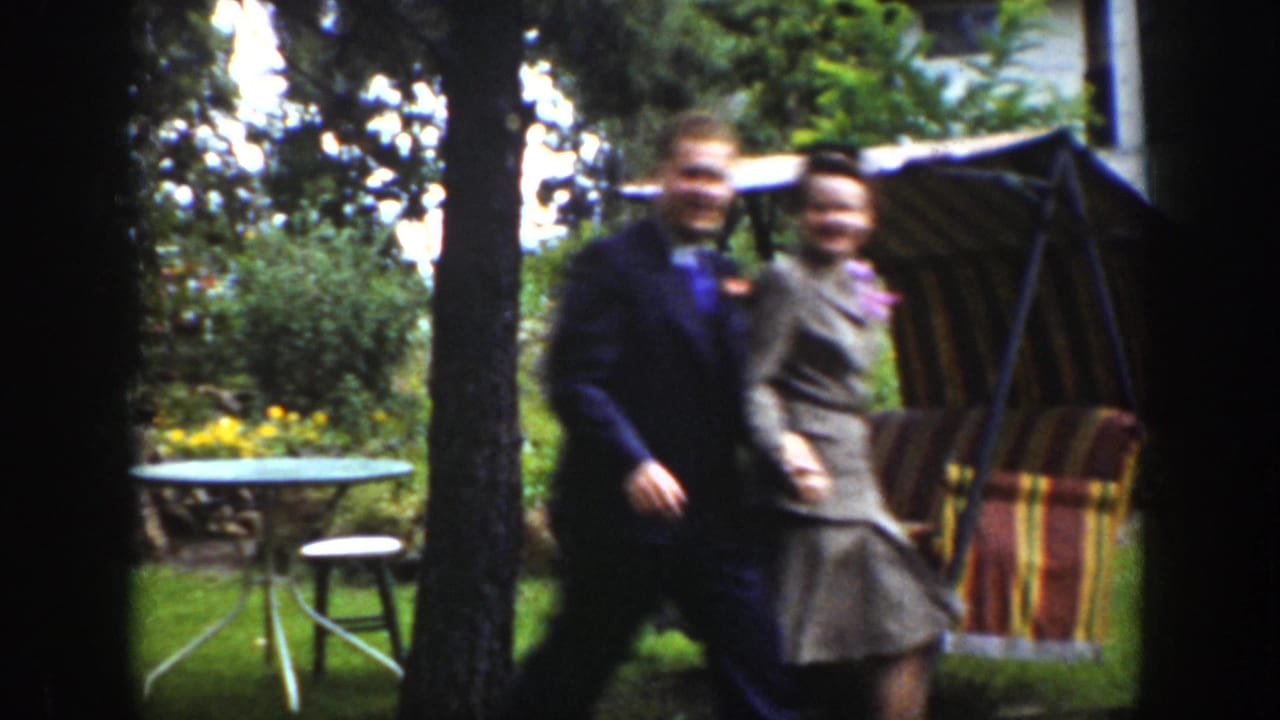 In a 1939 Wyoming home-movie frame, a well-dressed man and woman walk across a lawn beside trees and a striped patio swing.