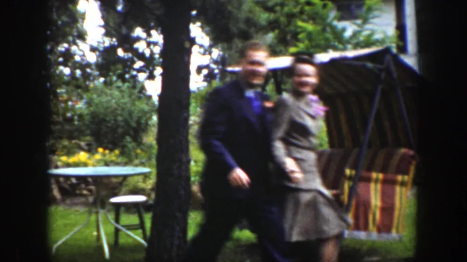 In a 1939 Wyoming home-movie frame, a well-dressed man and woman walk across a lawn beside trees and a striped patio swing.