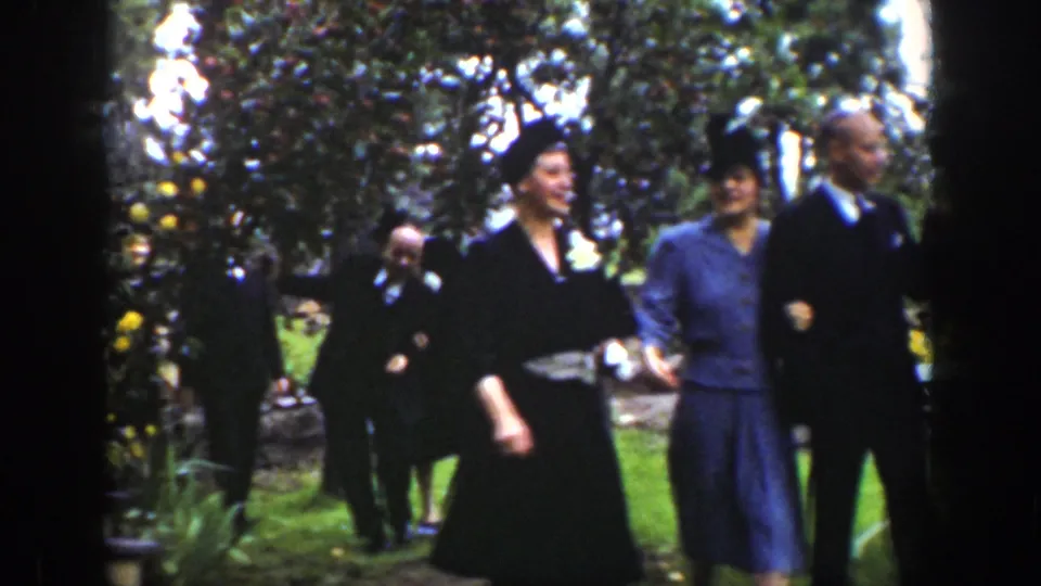 Guests in suits, dresses, and hats pass through a Wyoming garden scene, with trees and lawn framing the home-movie view.