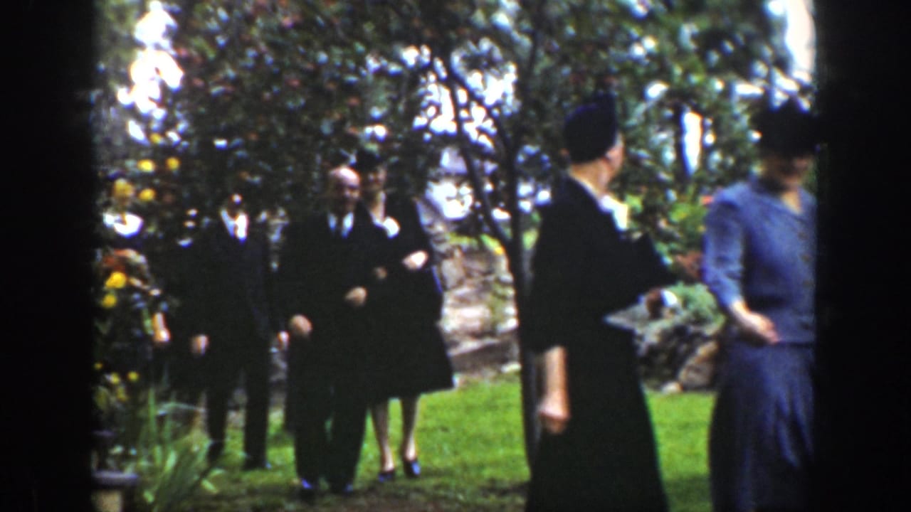 1939 Wyoming 8mm frame showing formally dressed adults walking through a leafy garden, with men in suits and women in dark dresses.