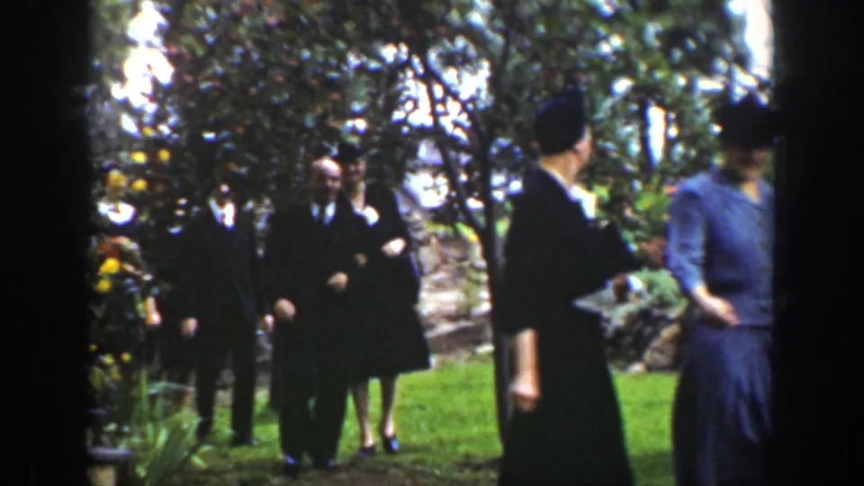1939 Wyoming 8mm frame showing formally dressed adults walking through a leafy garden, with men in suits and women in dark dresses.