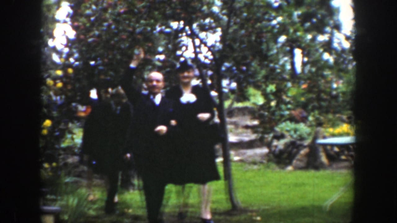 Two formally dressed adults walk arm in arm across a green garden lawn.