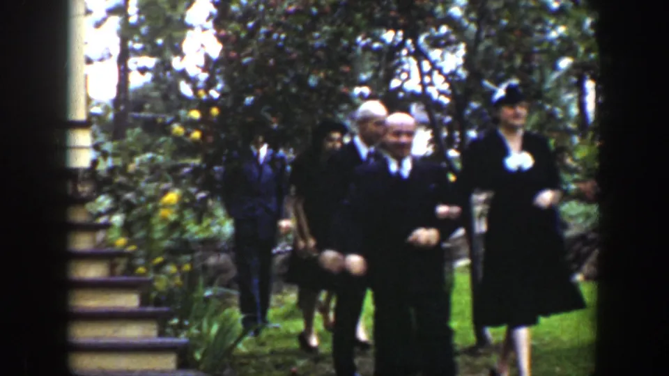 1939 Wyoming frame showing a small group of formally dressed adults walking together beside steps, trees, and garden plantings.