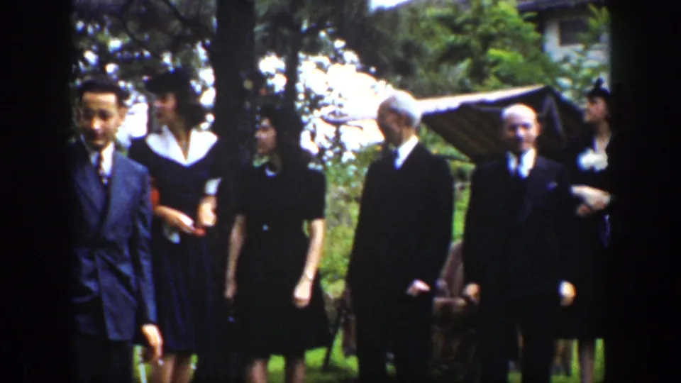 1939 Wyoming 8mm frame with a young suited man in front of adults in dark formal clothing beside trees and lawn.