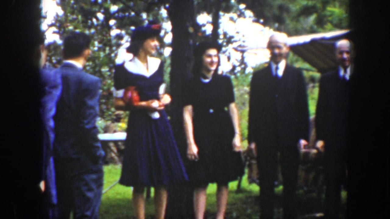 A man seen from behind faces a group of formally dressed adults standing near trees and a canopy-like shade.