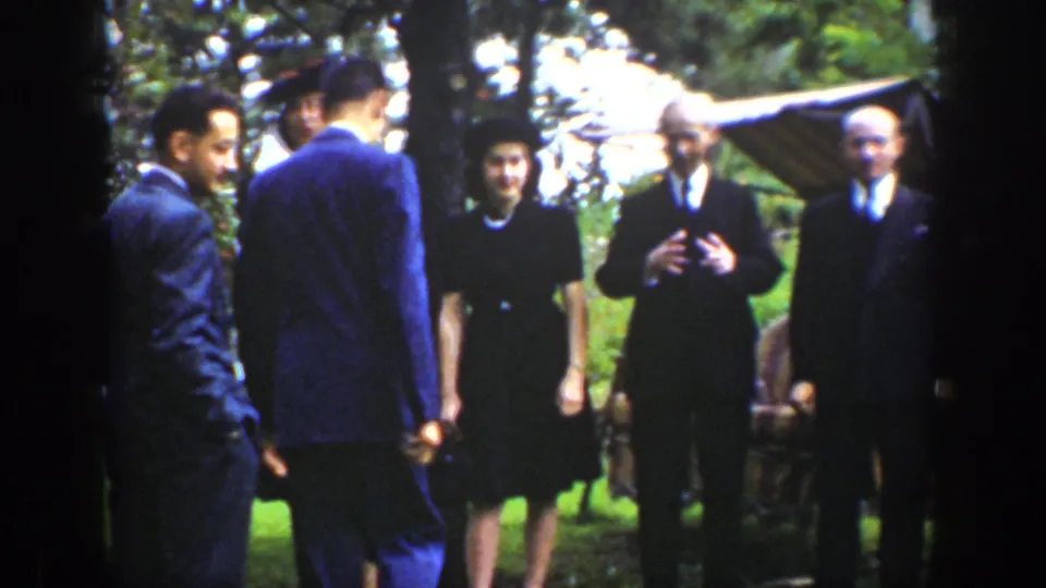 Adults in late-1930s formal clothing stand in a grassy outdoor setting, with two suited men turned away from the camera.