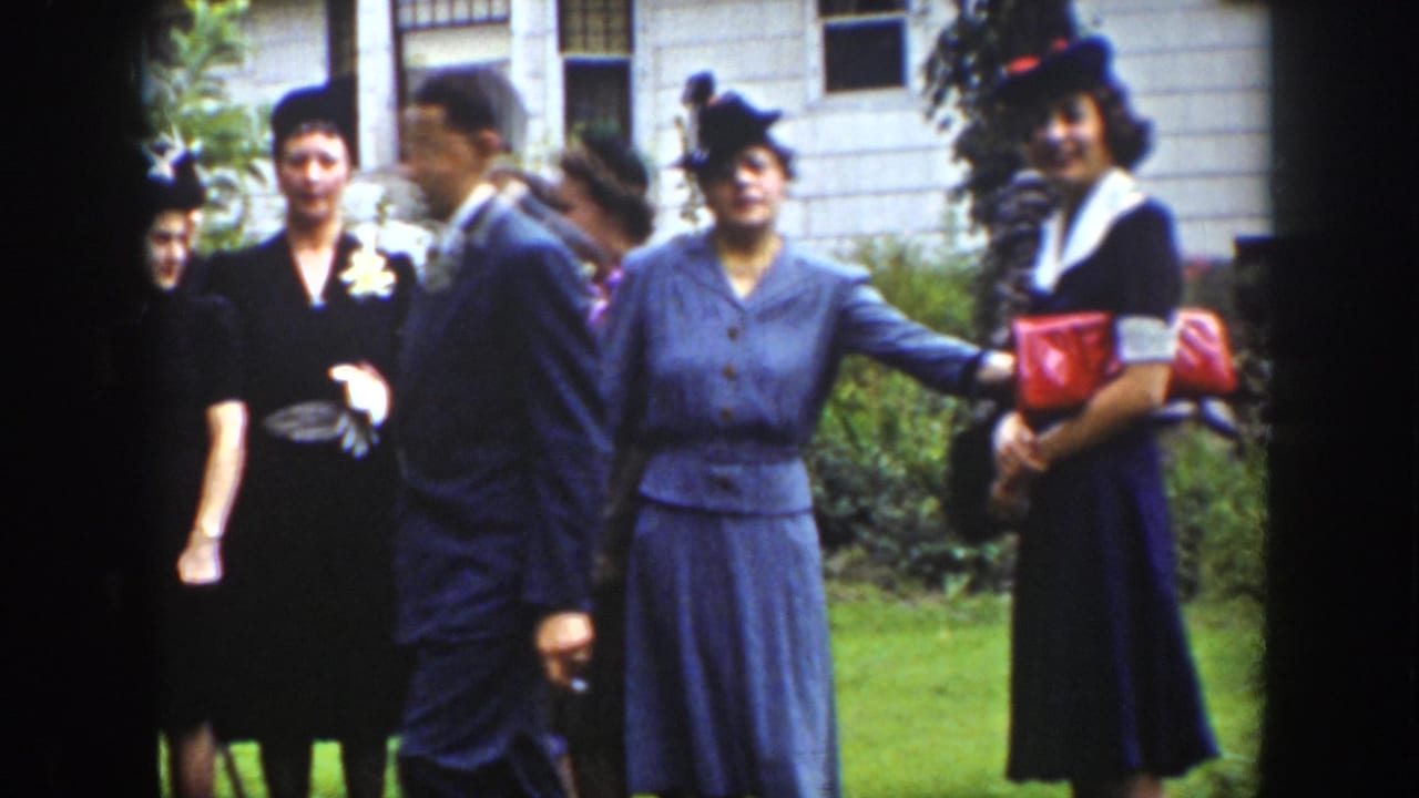 1939 Wyoming home-movie frame of formally dressed adults gathered on grass outside a white clapboard house during a wedding celebration.