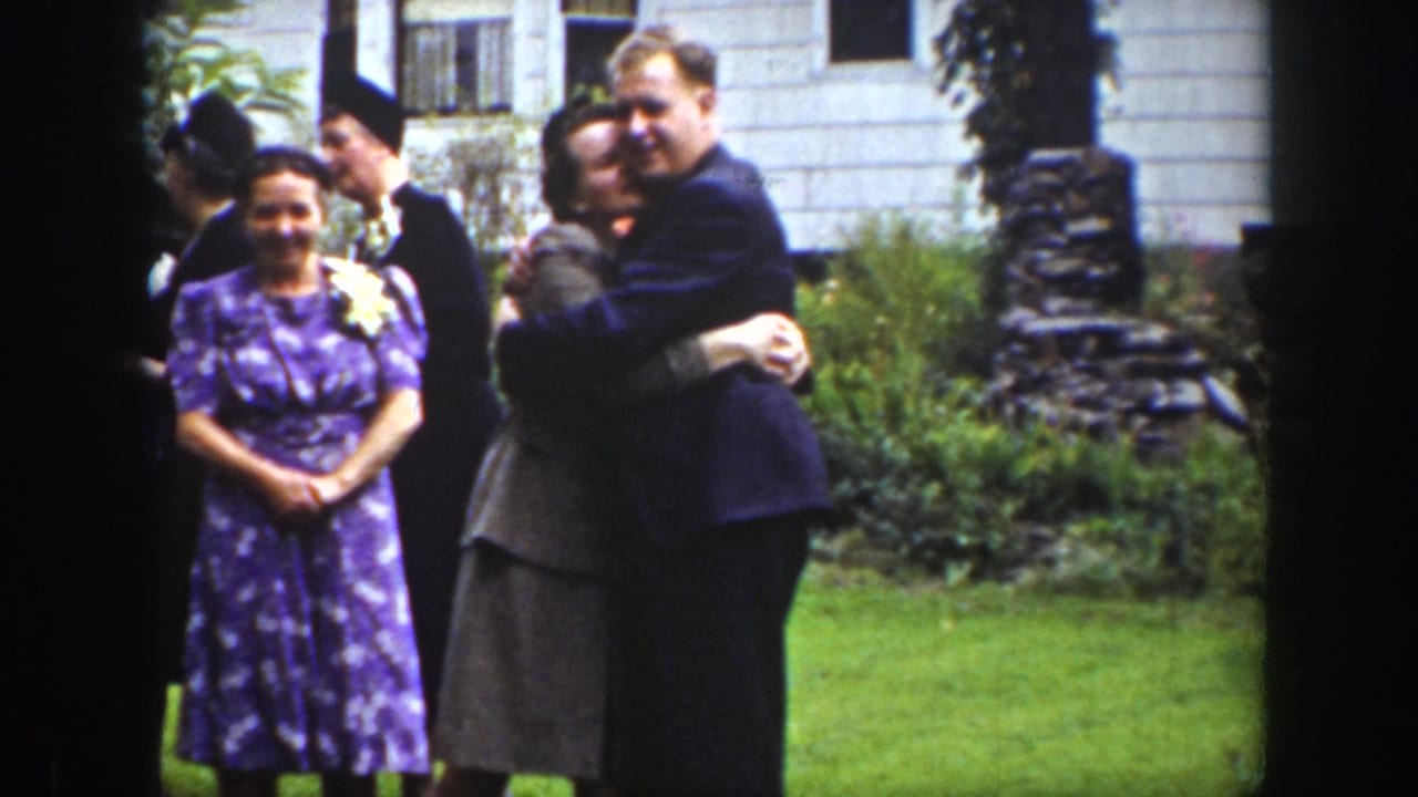 A smiling couple embraces while other wedding guests stand nearby on the lawn.