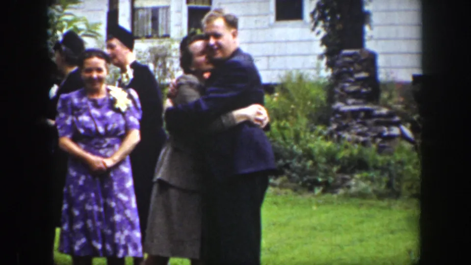 1939 Wyoming home-movie frame showing an adult couple embracing beside other formally dressed wedding guests outside a house.
