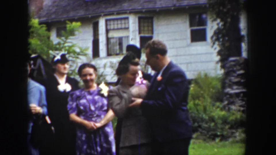 1939 Wyoming home-movie frame of wedding guests on a lawn, including a suited man facing a woman holding a small bundle.