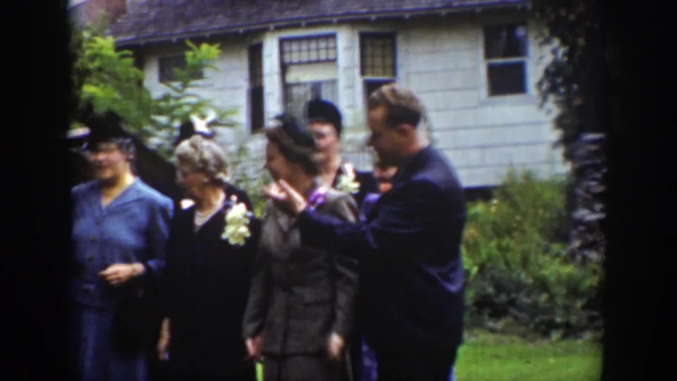 In Wyoming archival 8mm footage from 1939, adults in suits, dresses, hats, and corsages gather outside a house.
