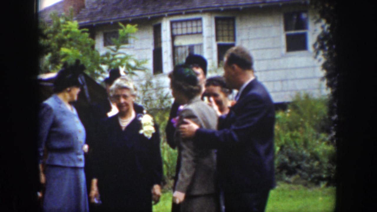 Adults in formal clothing adjust positions near a lawn and house.