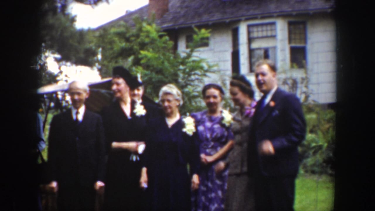 In 1939 Wyoming home-movie footage, adults in suits, hats, dresses, and corsages pose outdoors before a house.