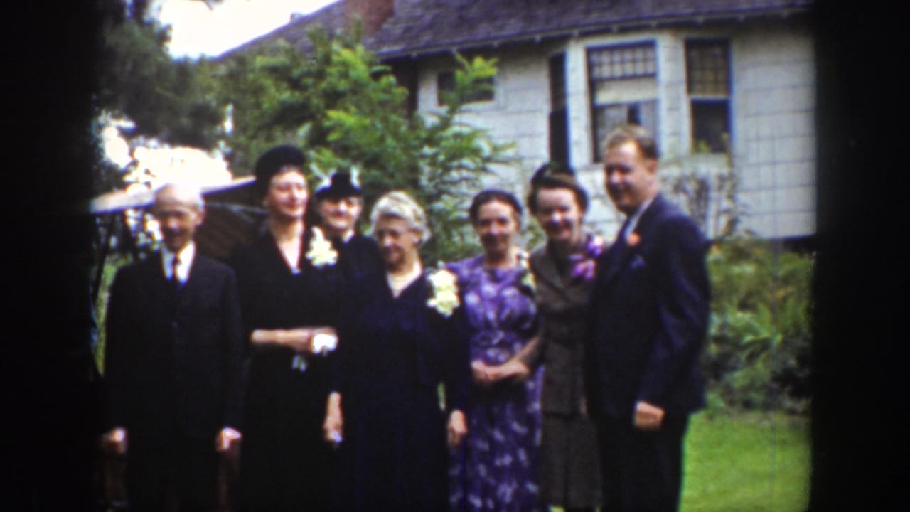 Seven adults pose in a line outdoors with a house and greenery behind them.