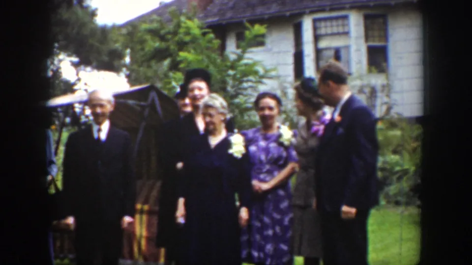 The home-movie frame shows the formal group adjusting their pose outdoors, with hats, suits, and dresses visible.