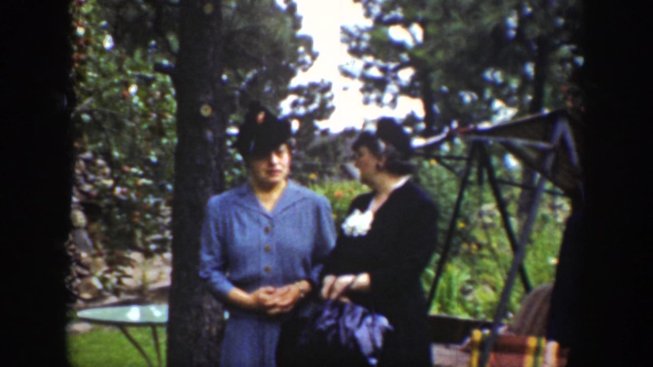 Two women in hats stand beside a tree and garden furniture.
