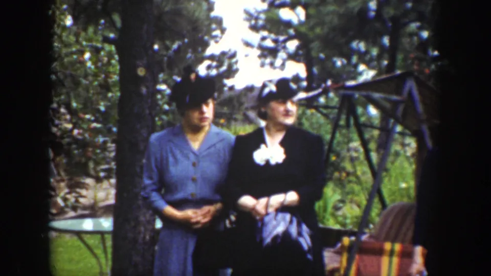 Wyoming 1939 home-movie frame showing two women in formal hats and corsages standing near a tree and outdoor seating.