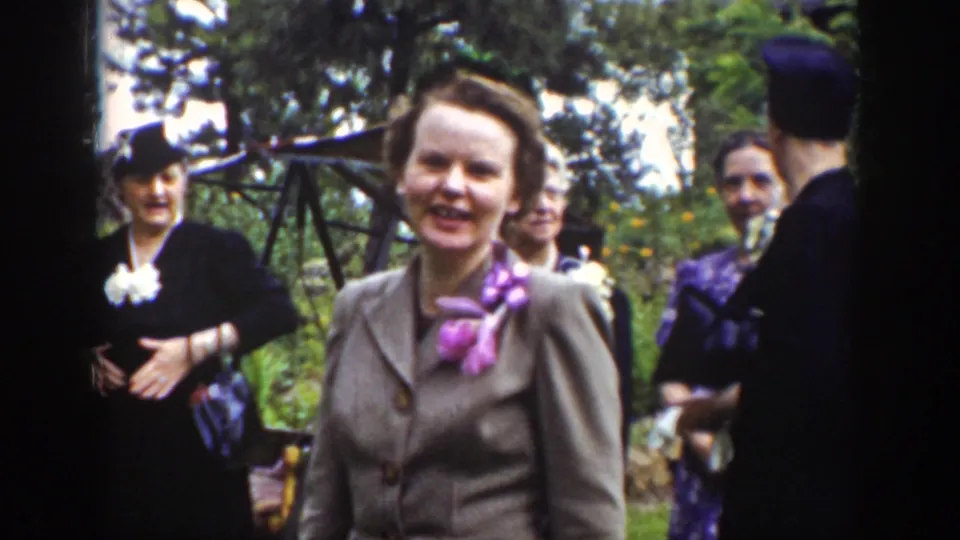 Wyoming 1939 frame with a smiling woman in a brown suit and pink neck scarf among formally dressed adults outdoors.