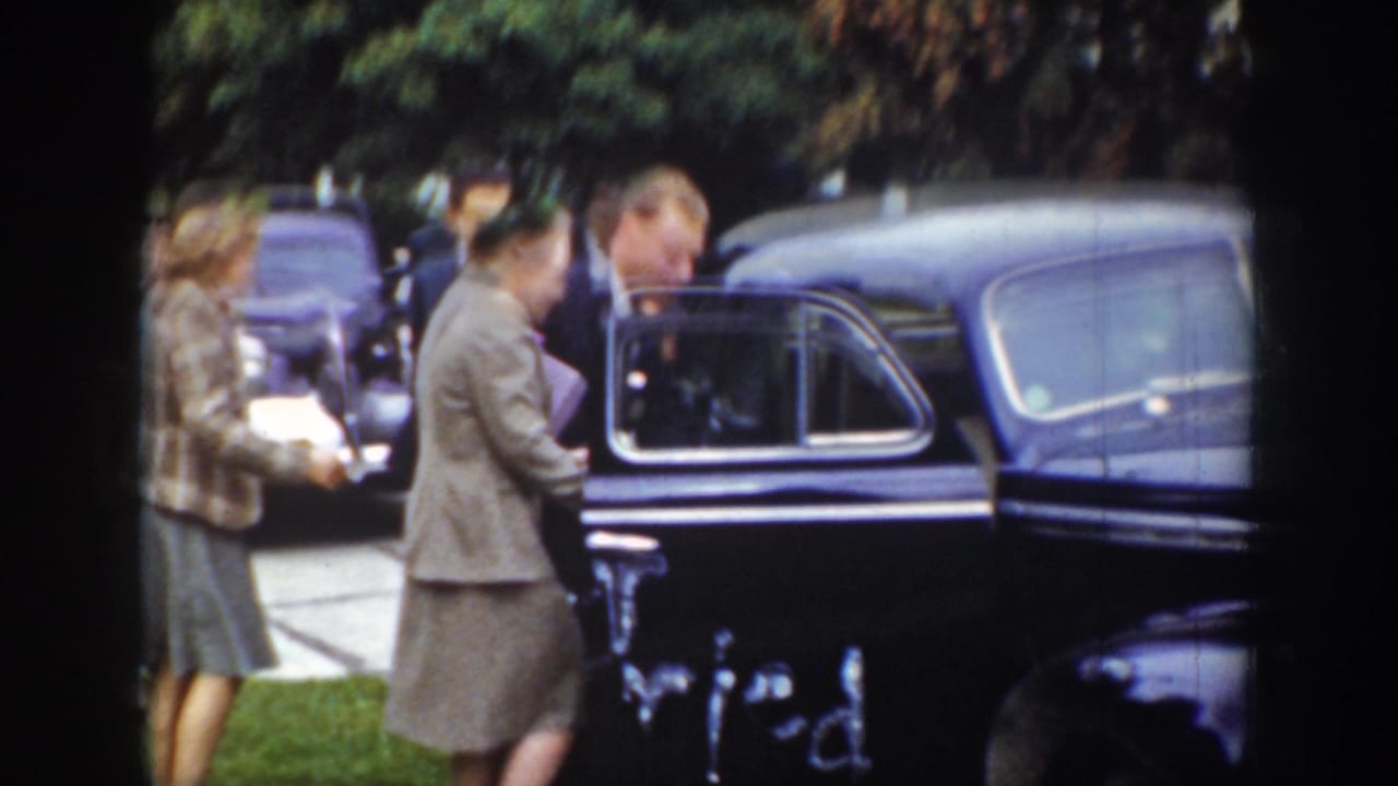 Adults in formal clothing stand beside a dark sedan with an open door in a grassy setting.