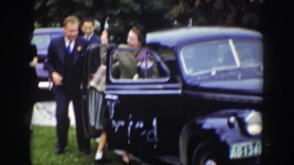 In the Wyoming 1939 footage, a woman appears to enter a dark sedan as suited men stand nearby in the grassy outdoor setting.