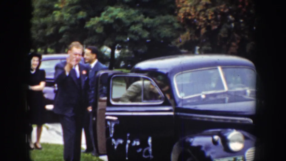A suited man raises his hand near a dark sedan, with another man and a woman visible behind him in the supplied 1939 Wyoming home movie.