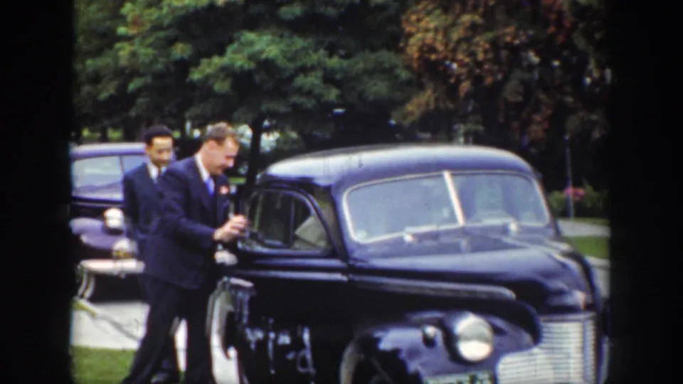A suited man reaches toward the dark sedan door while another man stands behind him, with trees and another vehicle in the background.