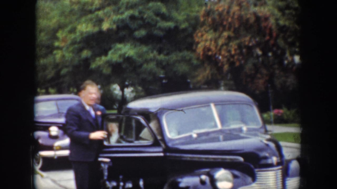 In 1939 Wyoming home-movie footage, a suited man stands beside a dark prewar sedan parked along a tree-lined street.