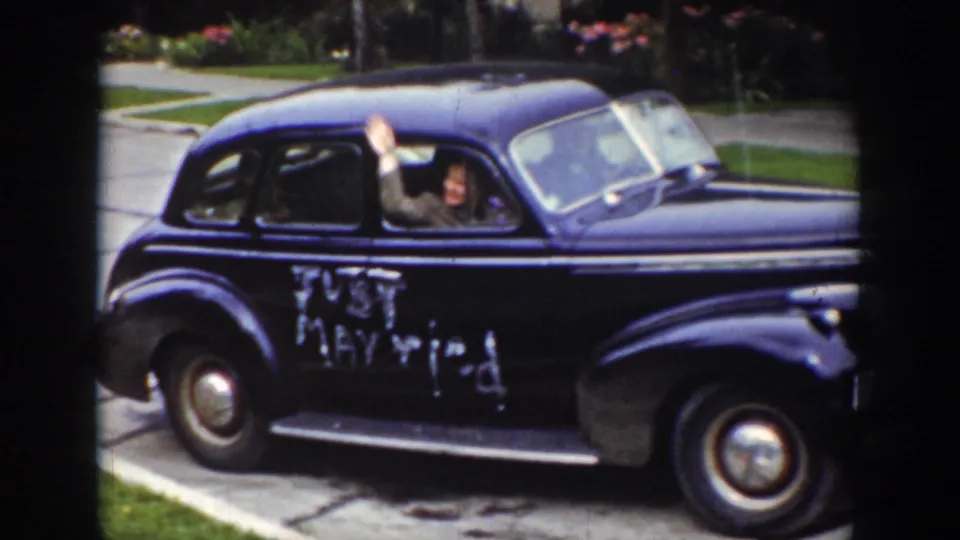 A passenger waves from the rear window of a dark sedan with Just Married written in white on its side.