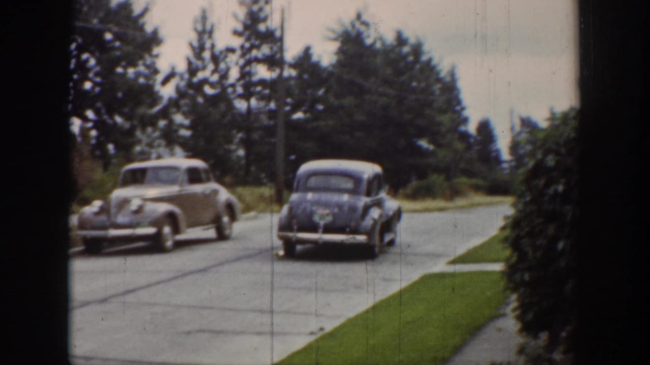 1939 Wyoming home-movie frame showing two period cars on a quiet tree-lined street with lawn edges and utility poles.