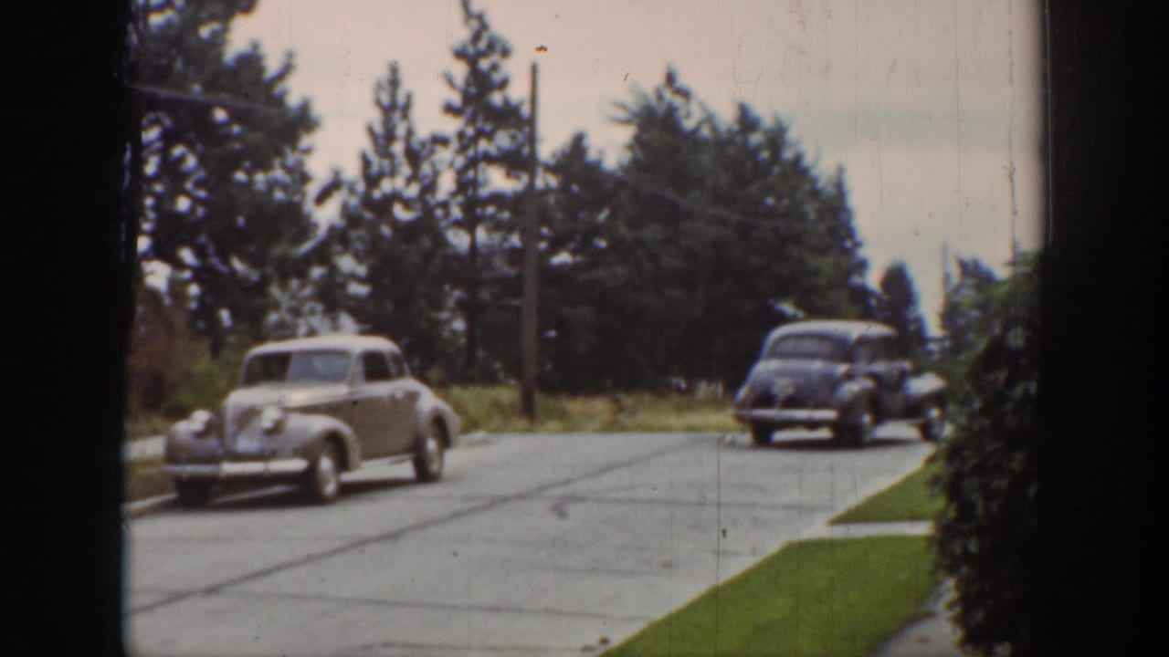 A light-colored car passes while a darker car follows on the same street.