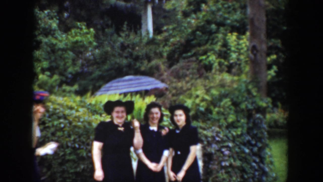 Three women in dark dresses stand under a striped umbrella in a green garden.