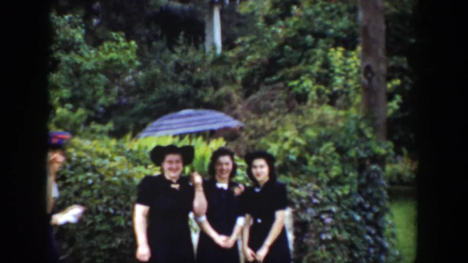 In 1939 Wyoming home-movie footage, three women in dark dresses and hats smile beneath a striped umbrella in a lush garden.